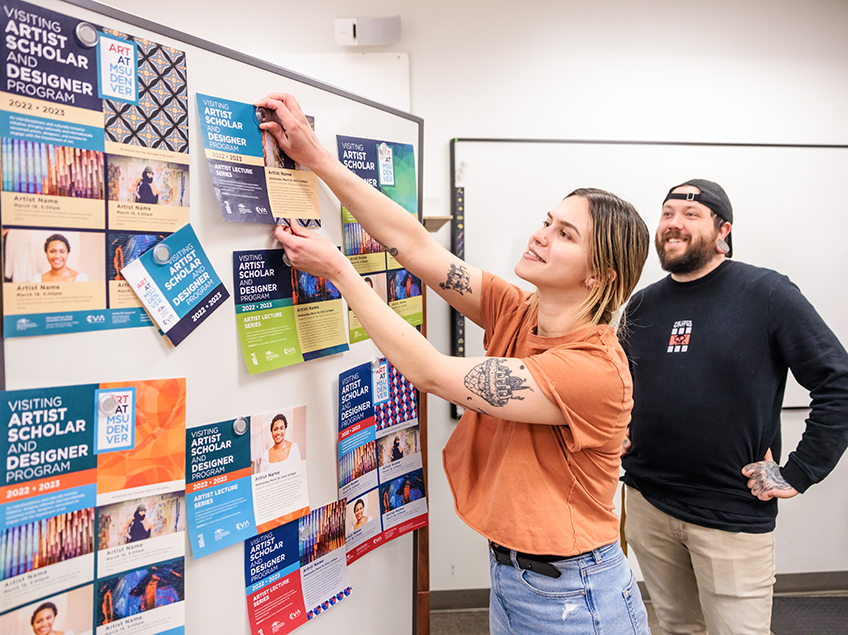 Students post finalized examples on a white board, for their project which creates advertisements for the Visiting Artist Scholar and Designer Program. In use are many bright and complementary color palettes including teal, sky blue, orange, lime green and navy, along with a dark cream and light lavender. Both students happen to have prominent tattoos showing on their arms.