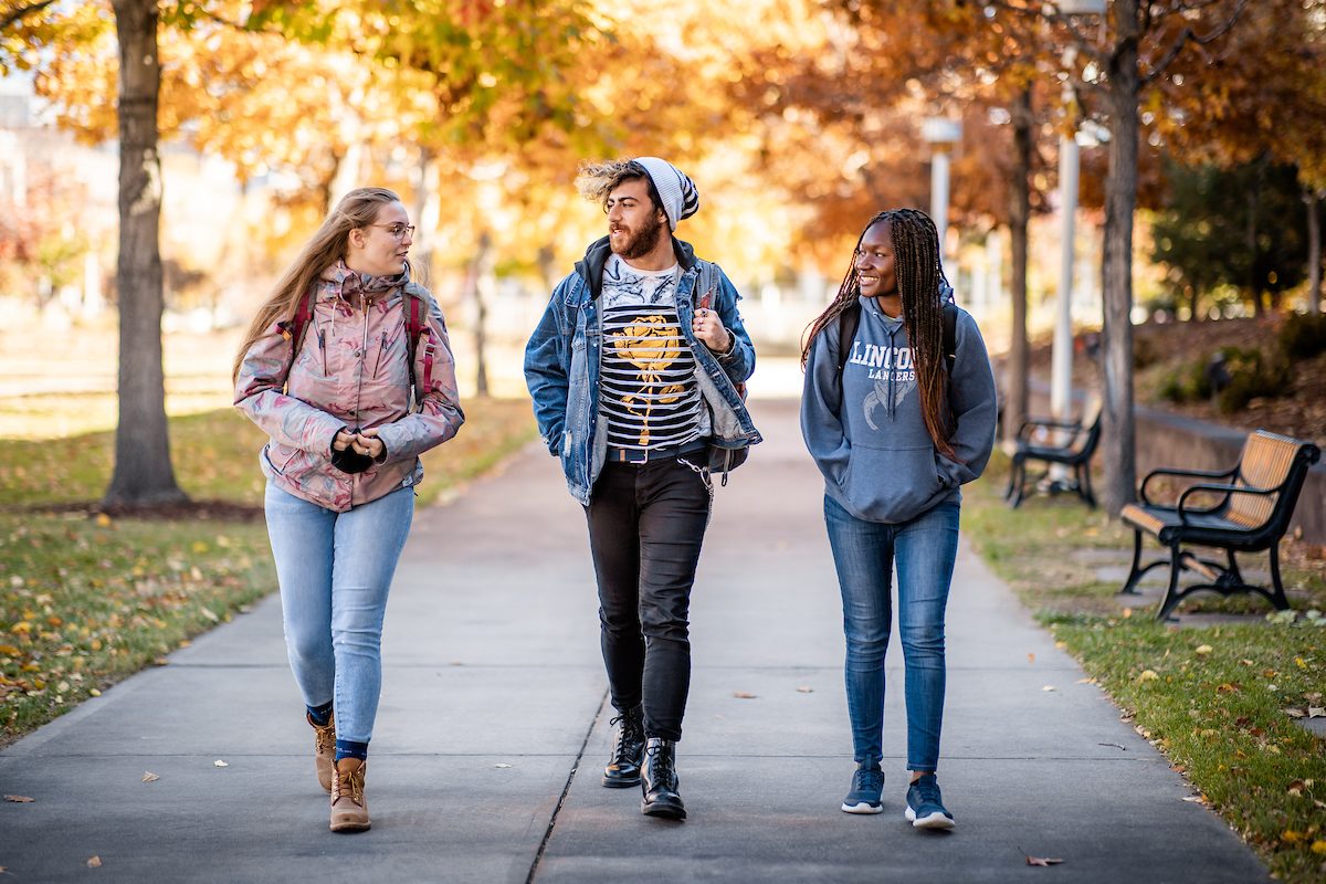 Three students walking on a tree-lined campus pathway during the fall. The student on the left is wearing a pink jacket and glasses, the student in the middle is wearing a denim jacket and beanie, and the student on the right is wearing a gray hoodie with 'Lincoln Lancers' written on it. The ground is covered with fallen leaves, and the trees in the background have autumn foliage.