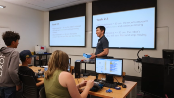 An instructor in front of projector screens holding a robotics kit, teaching teenagers.