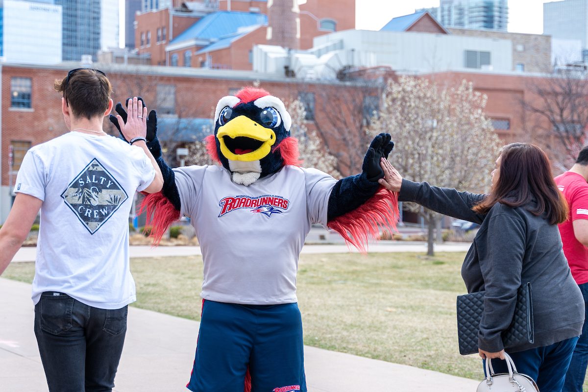 Rowdy the Roadrunner, MSU Denver's mascot, giving high-fives to a student and an adult during an outdoor event. The student is wearing a white 'Salty Crew' T-shirt, and the adult is wearing a gray jacket and carrying a black quilted bag. The background features buildings and trees on the MSU Denver campus.