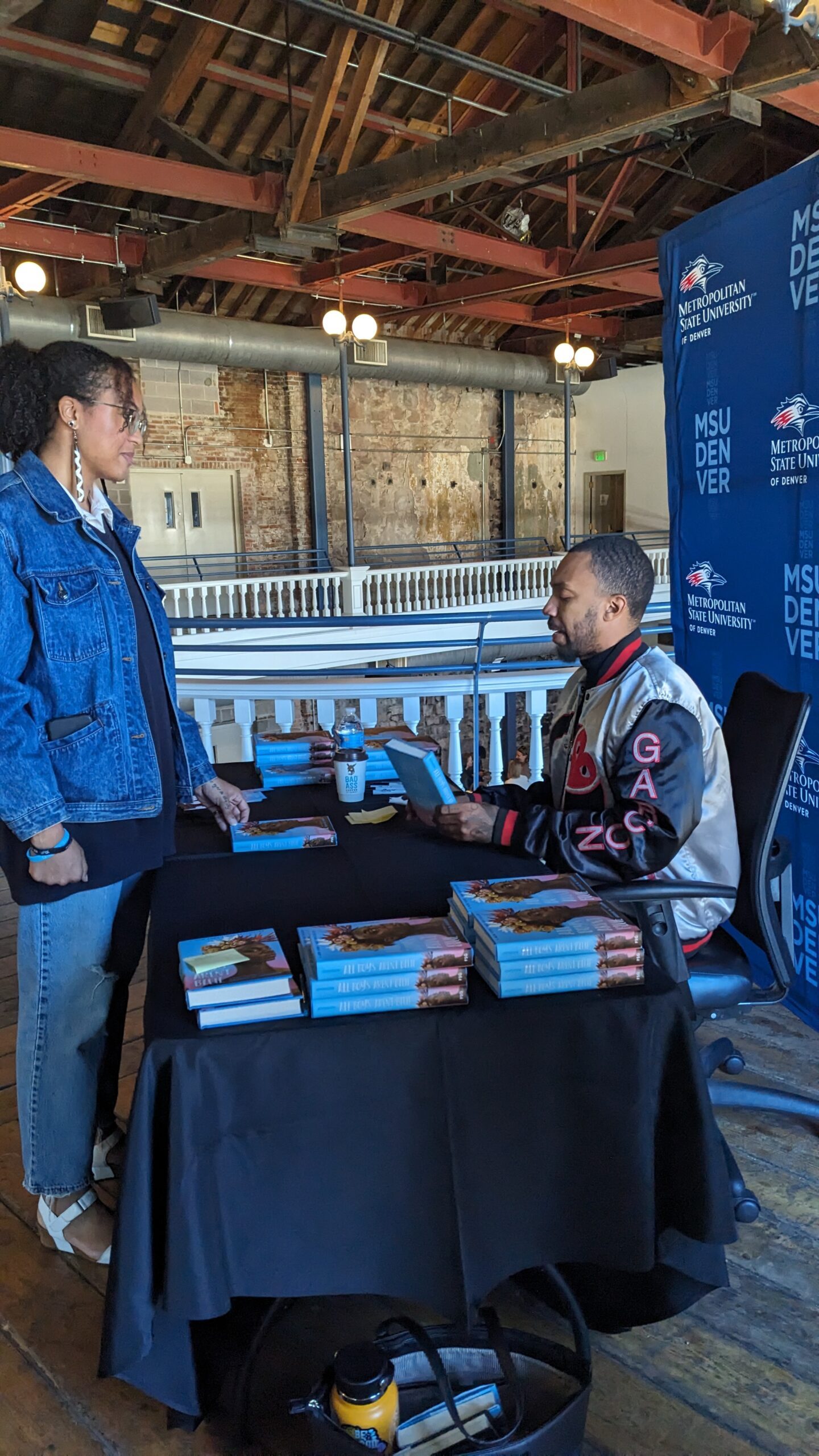 African-American author sits at a table with stacks of books speaking across the table with a standing student. Student is waiting to get a copy of a book signed by the author