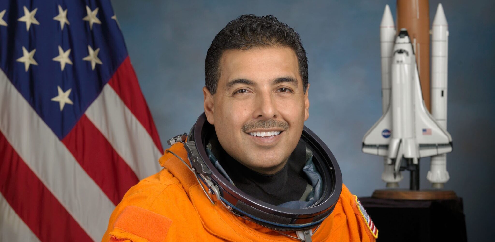 Astronaut Jose M. Hernandez, mission specialist in Orange space suit, American flag and Space shuttle model in the background