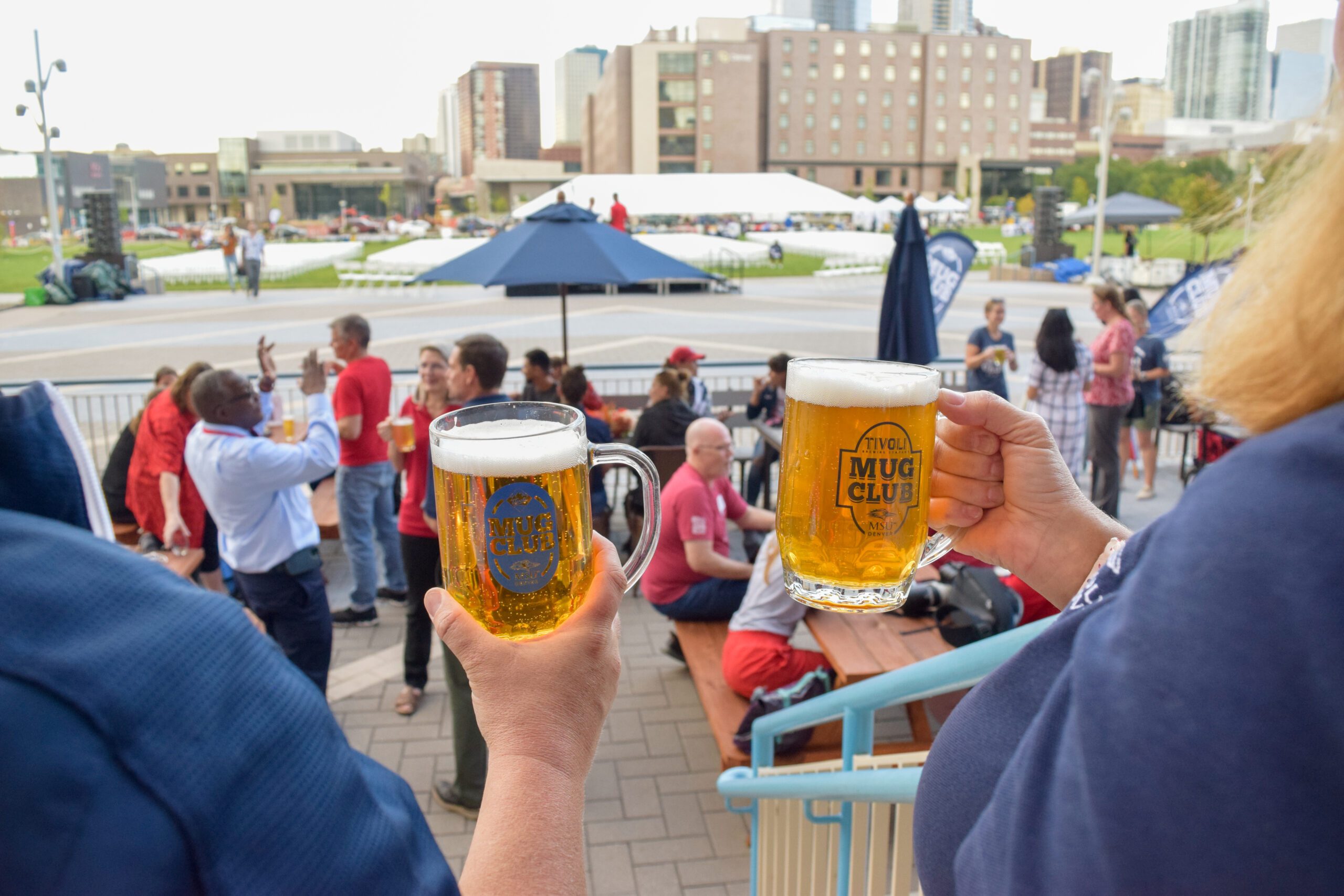 Two members of the MSU Denver community holding up their Mug Club glasses in front of a group of people enjoying Happy Hour on the Tivoli Tap House porch.