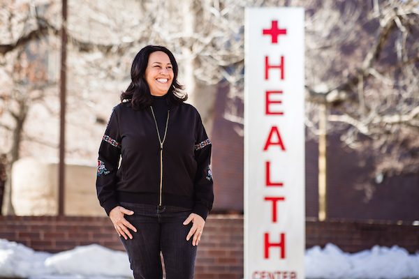 Maria Freyta stands in front of the Health Center at Auraria in a black jacket. The sign behind her reads 'Health'