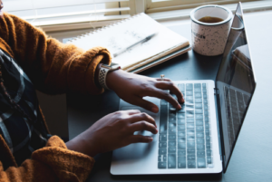 Overhead image of hands typing on a laptop with coffee and notebook nearby.