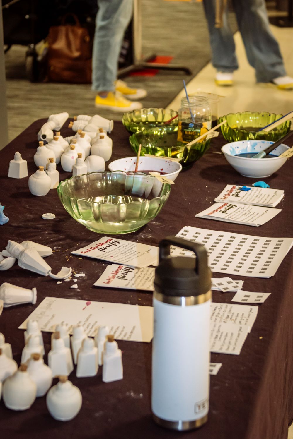 Colored bowls with paintbrushes and paint water, white water bottle, white ceramics and paper sitting on a table