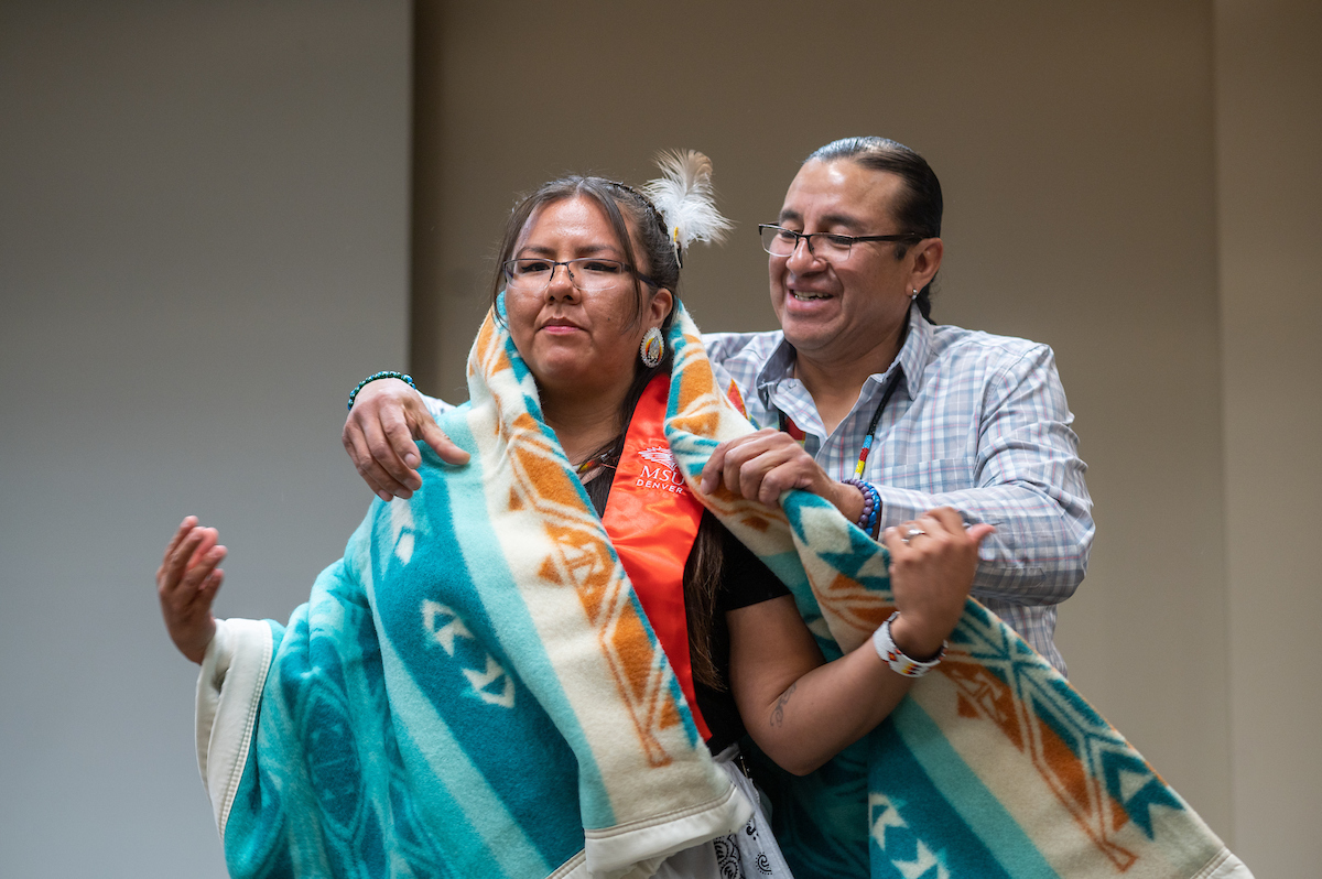 An older person wrapping a ceremonial Native-Indigenous blanket around a graduating MSU Denver student