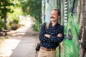 Hamilton Nickoloff, a young man with his haired pulled back leans against a colorful wall mural, smiling