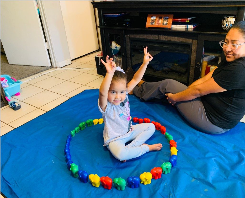 A parent and a child during a home visit, the young child with their arms up inside a ring of colorful elephant toys