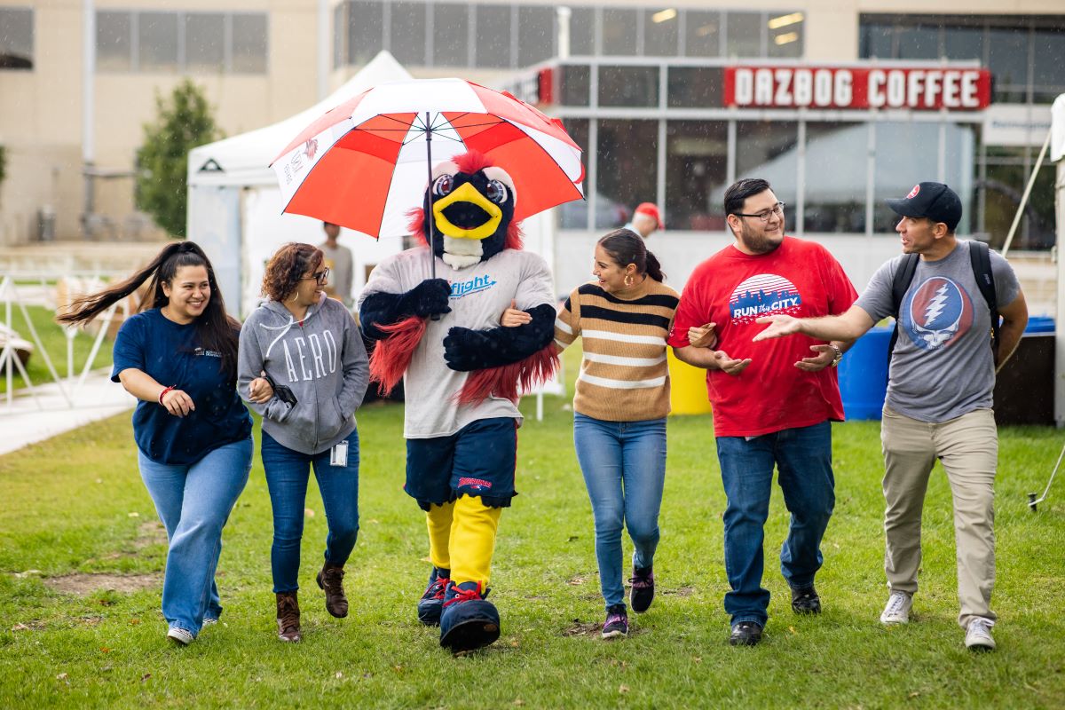 Five students walk in the rain with their arms linked with Rowdy the mascot in the middle holding a red and white striped umbrella