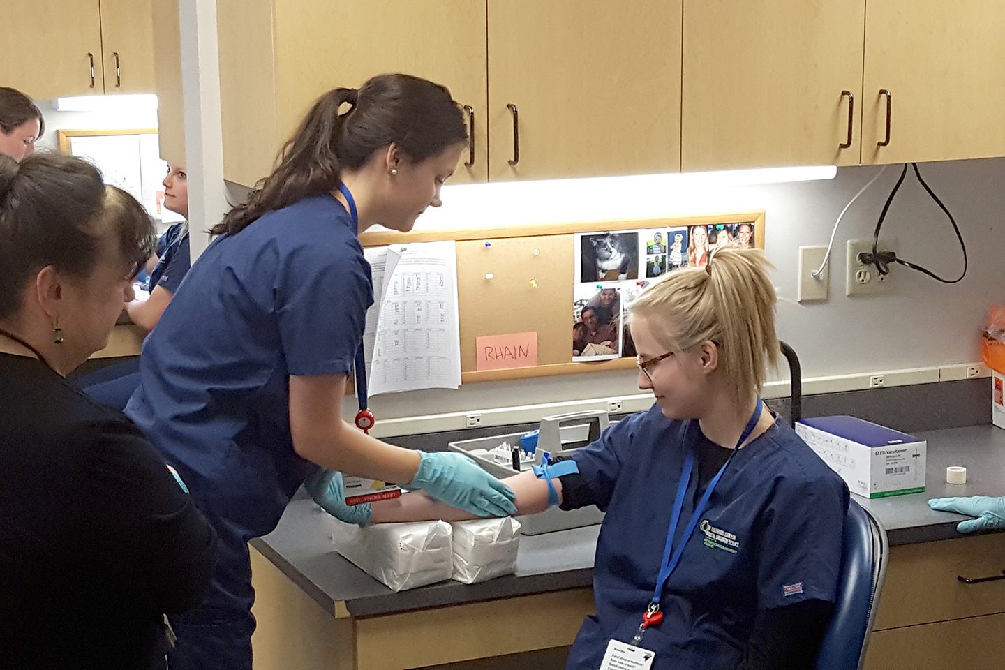 Female student drawing blood from another female student with instructor watching