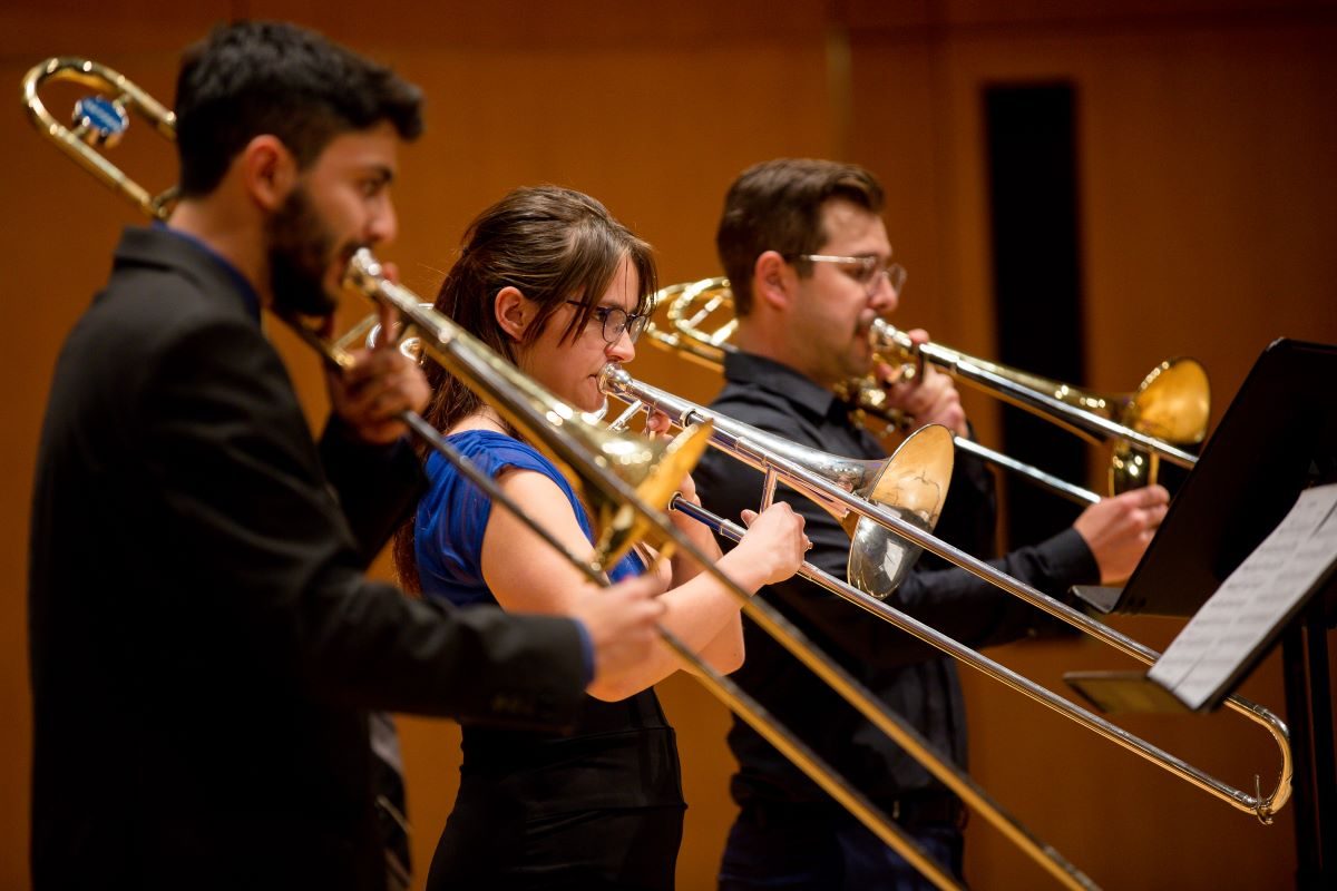 Three students read music sheets while playing the trombone