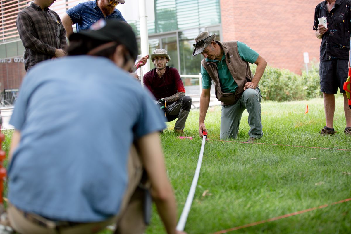 A MSU Denver professor and students kneeling and measuring a grassy area with a tape measure and red string