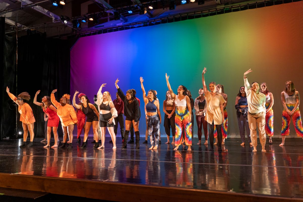 Students in costume hold one hand in the air as they bow after a performance in front of a rainbow backdrop