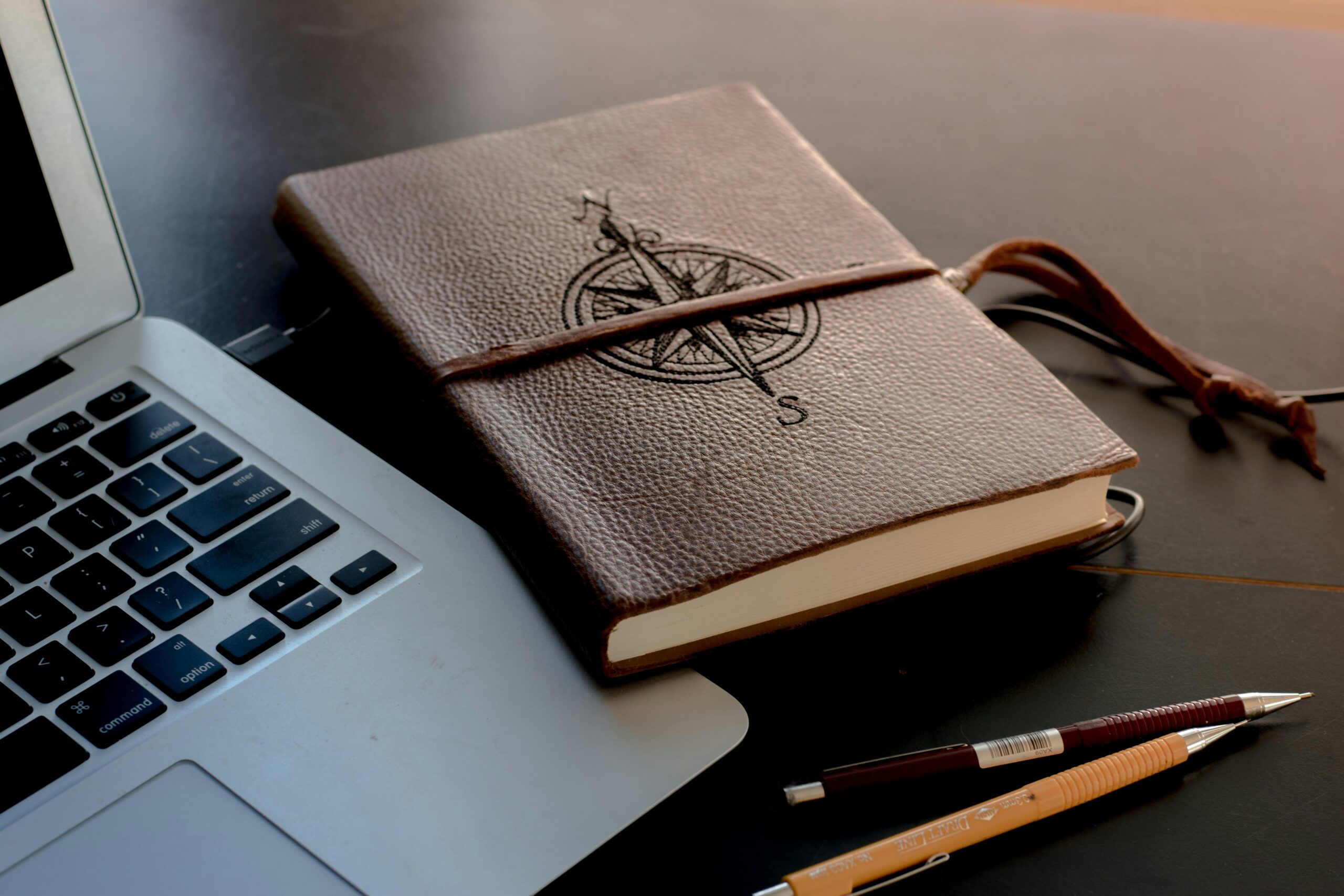 Photo composed of a laptop computer, two mechanical pencils, and a brown leather notebook with a compass rose image stamped into the cover with a strap over the middle of the notebook. On a brown table top.