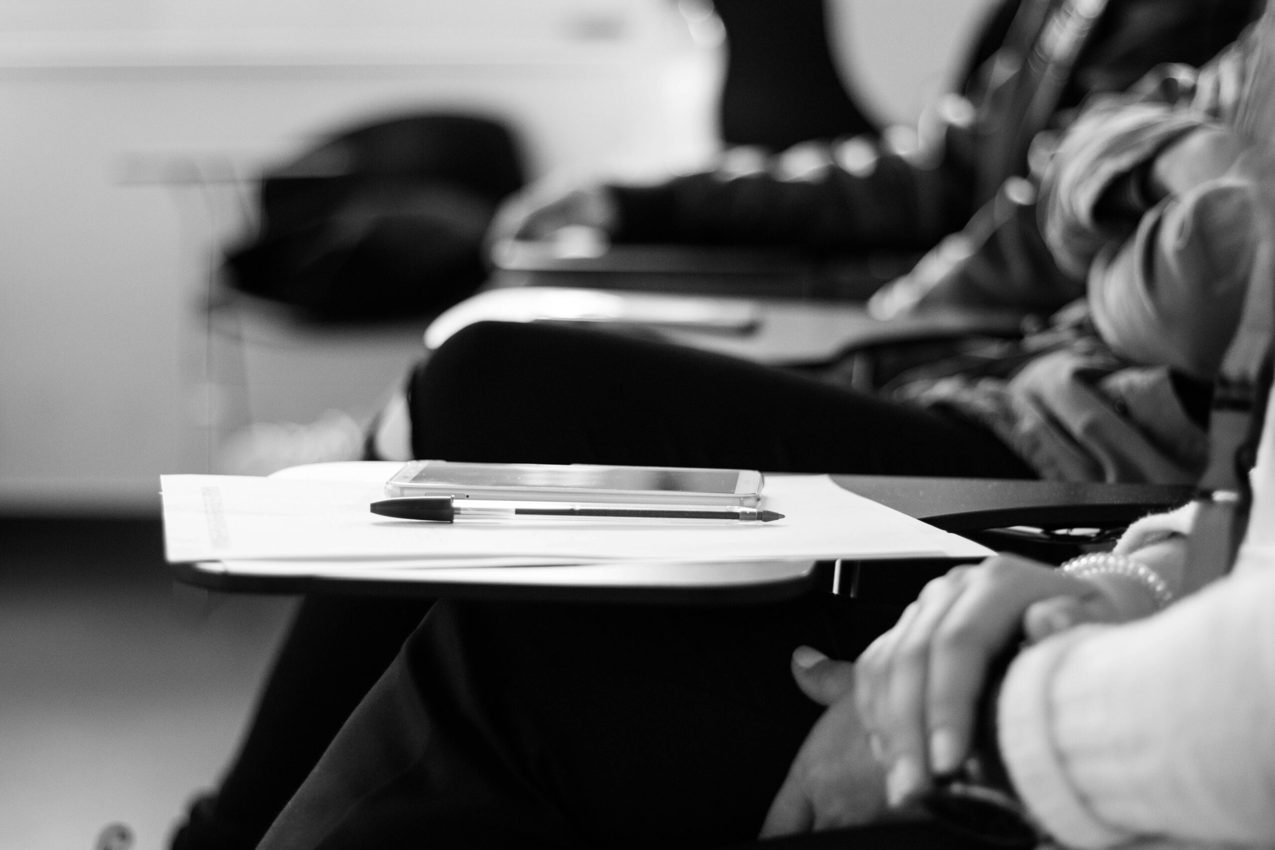 Black and white photograph of an individual at a school desk with paper, pen, and smart device on the desk